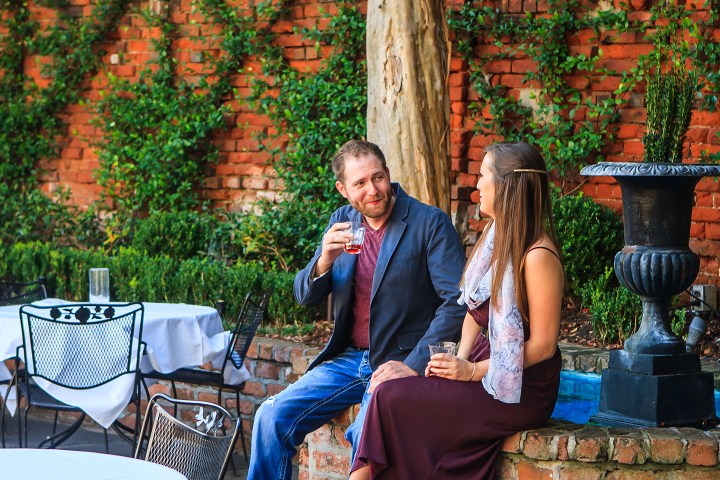 Two people seated outdoors, drinking by a brick wall and fountain.