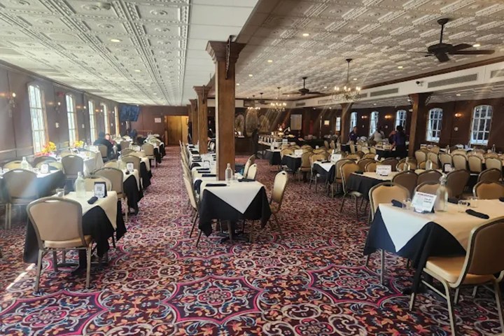 Spacious dining room with ornate carpet, set tables, chandeliers, and large windows.