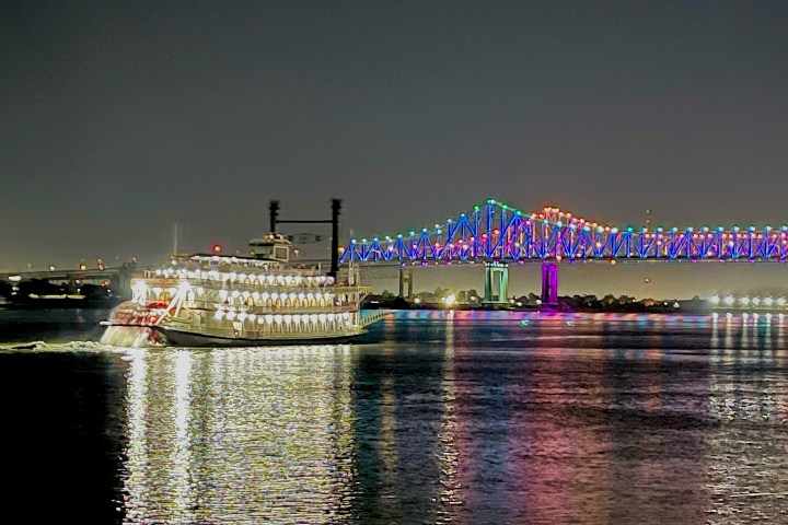 Lit riverboat cruises near a colorful illuminated bridge at night.