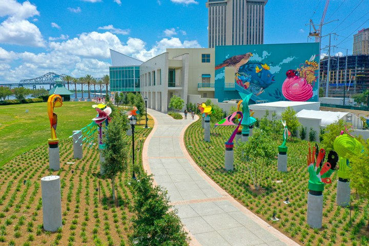 Colorful sculptures line a walkway outside a modern building with a mural, under a bright blue sky.