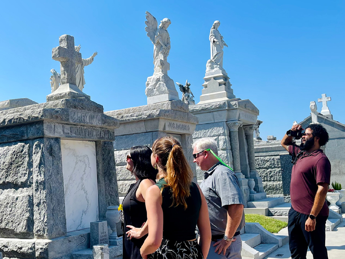 Group of people walking through a historic cemetery with ornate mausoleums on a sunny day.