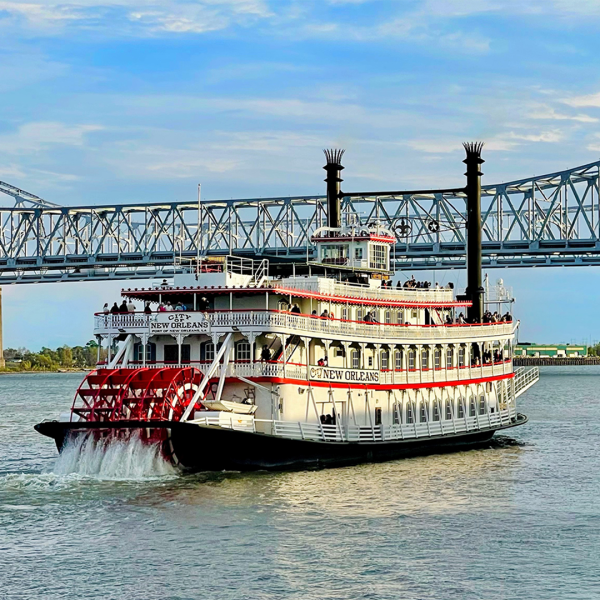 Steamboat on a river with a bridge in the background under a blue sky.