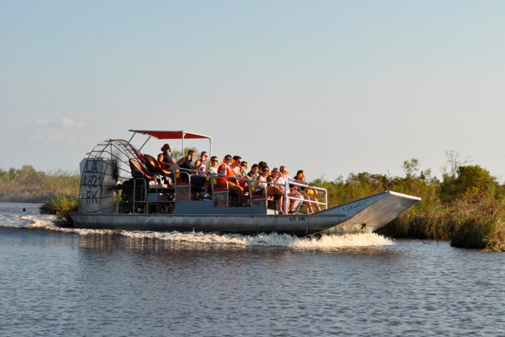 Tourists ride on an airboat through a marshy waterway under a clear blue sky.